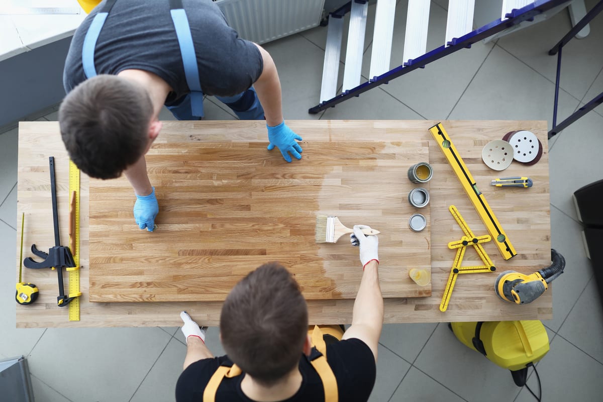 Overhead view of two refinishers coating a wood counter surface with tools laid out beside them